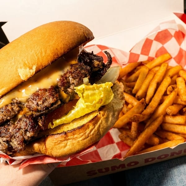 Hand holding a double cheeseburger with lettuce and sauce, next to a box of fries.