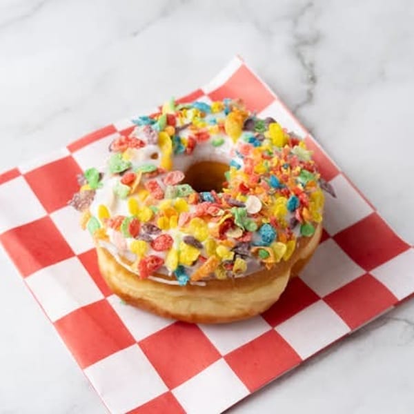 A white-frosted donut with colorful cereal sprinkles sits on a red and white checkered napkin.